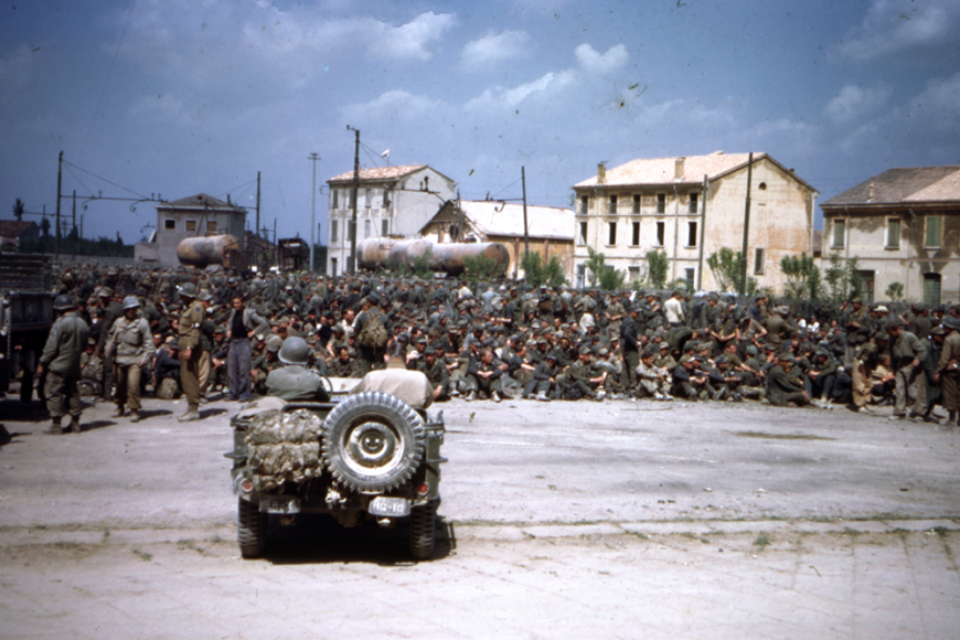 A very large group of soldiers sits in the center of Camp Hale consisting of large buildings, sparse trees, and gas tanks. Two people in a small army vehicle face the group of soldiers.