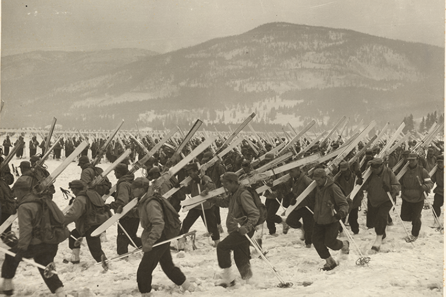 A large group of warmly dressed soldiers carrying skis and poles are trudging across a snowbank in Camp Hale, with mountain ranges in the background.
