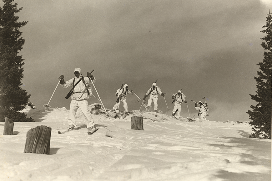 Five soldiers in full gear, skiing over a snowy crest through a couple of trees and stumps.