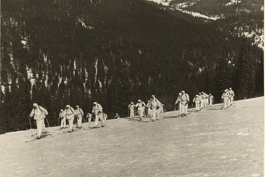 A group of many soldiers in white snow gear are cross country skiing over a snowbank, with large tree-covered mountain in the background.