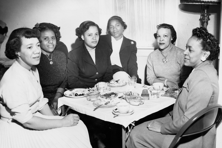 A black and white photo of six African American women sitting around a small table set with teapots, plates, and cups.