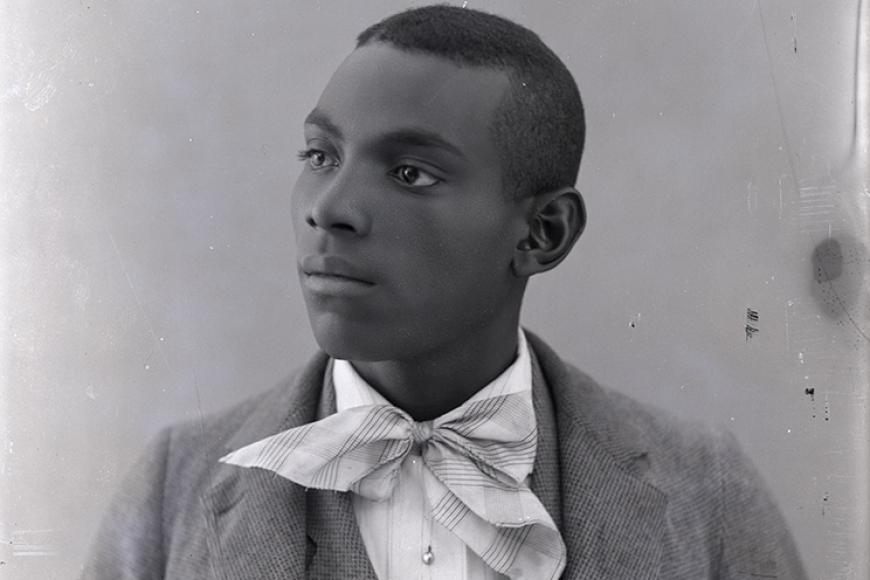 The image depicts a studio portrait of a young man. Only his bust and head are visible in the portrait. He is posed with his head looking to the left. The young man wears a lightly colored checkered three-piece suit with a white pin-stripe button down shirt. He also has on a plaid neckerchief tied into a bow.