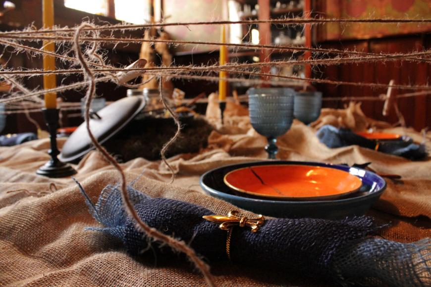 Closeup of dining table at 12 tablecloths art exhibition. There is a burlap tablecloth, burlap napkins, and burlap twip hanging above. There's a golden Fleur de Lis around the napkin. An orange ceramic plate sits on a blue ceramic charger.
