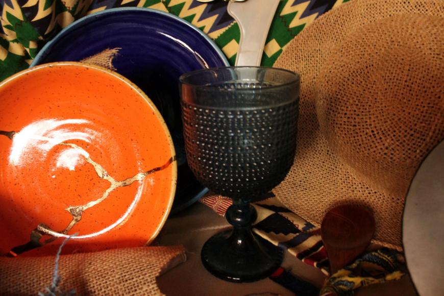 A clear, textured goblet sits in front of a star hat, a woodne sppon, and blue and orange ceramic plates. The orange plate has a golden crack through it. Behind these items is a green, yellow, purple, and black textile with African patterns.