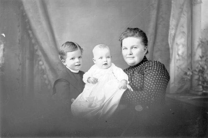 Studio portrait of a woman, a young boy and a baby. The baby wears a light colored gown. The boy has a part in his hair and wears a dark colored jacket. The woman wears a polka-dot blouse and her hair is pulled back into a pompadour.