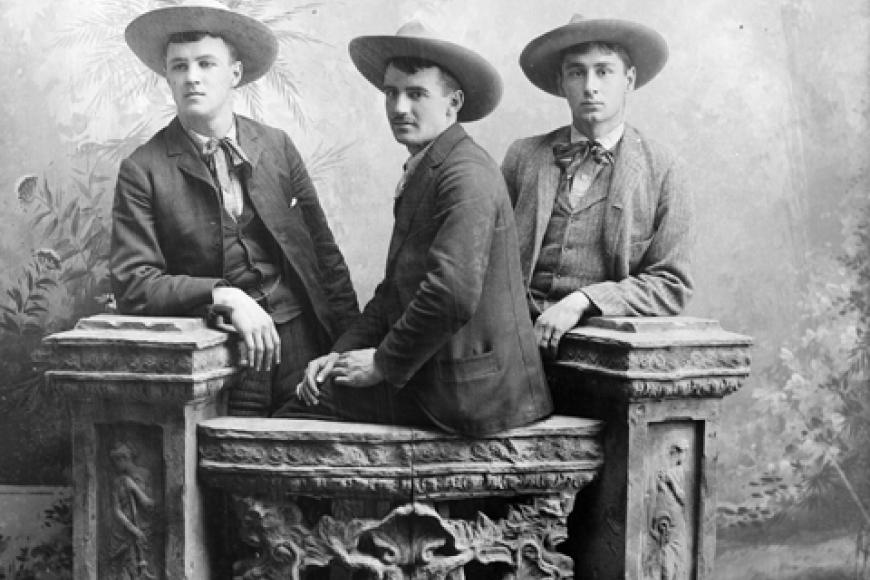 Studio portrait of a group of men. They pose on and near a decorative plaster banister. They wear suits, ties, and wide-brimmed hats.