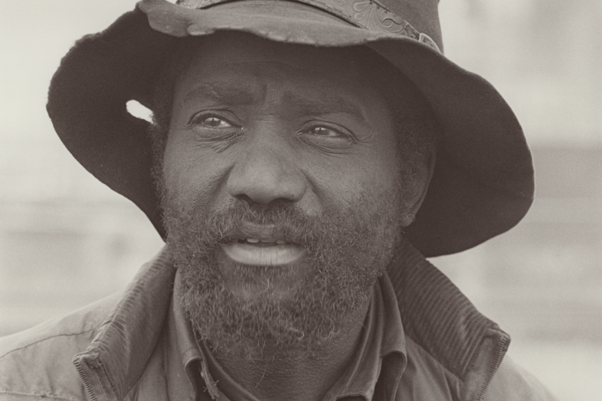 A portrait, showing Leonder Taylor from the chest up and looking just off camera, wearing a worn brimmed hat and jacket. 