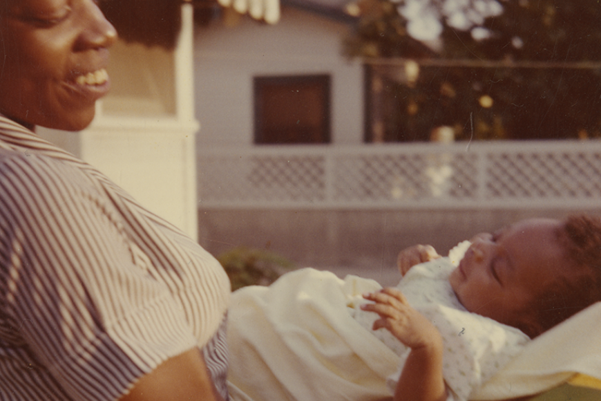 A woman (Alice) stands in a suburban backyard and smiles lovingly while cradling her baby (Rich) in her arms. They are both wearing white blouses.