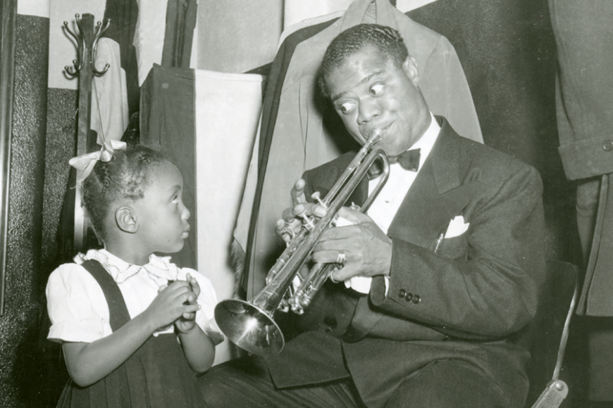 A black and white photograph of Louis Armstrong sitting and playing his trumpet for a little girl.