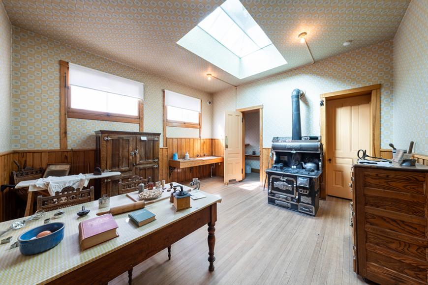 Interior of the Center for Colorado Women's History kitchen. There is an antique stove, a table with artifacts, and a sun light.