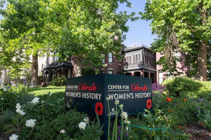 Lush, green exterior of the Center for Colorado women's history. The sign for the museum sits in front of the purple brick house, surrounded by trees.