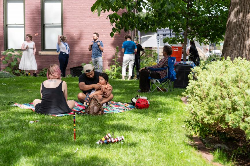 A family sits on the lawn of the center for Colorado Women's History. Other guests socialize in the gardens behind them.