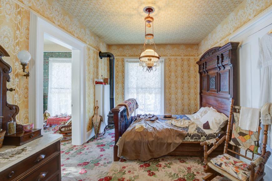 A Victorian furnished bedroom with a floral carpet, yellow wallpaper and a large bed with an antique wooden headboard.