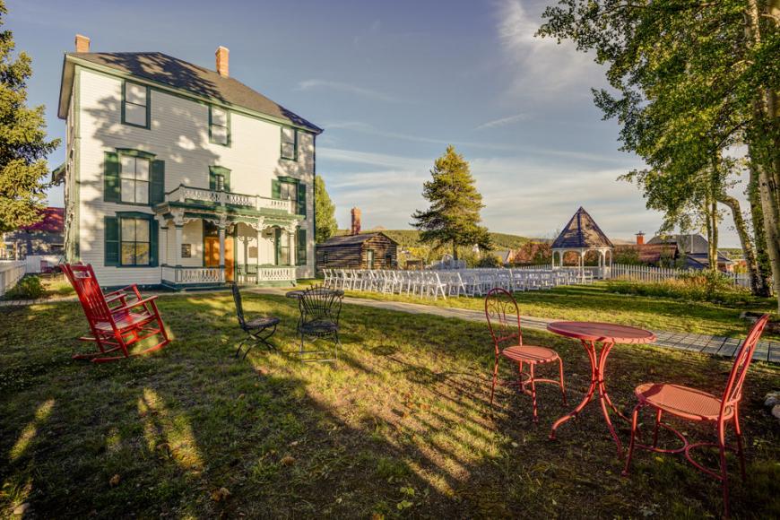 Large historic three-story home with white siding and dark green shutters and trim on a grassy lawn surrounded by trees.