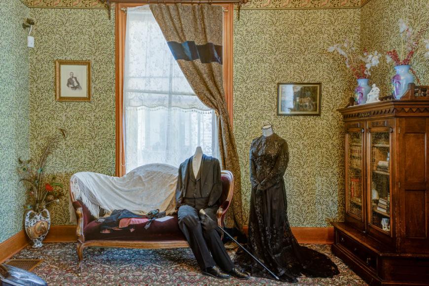 A Victorian era furnished room with two mannequins dressed in black attire on a velvet sofa.