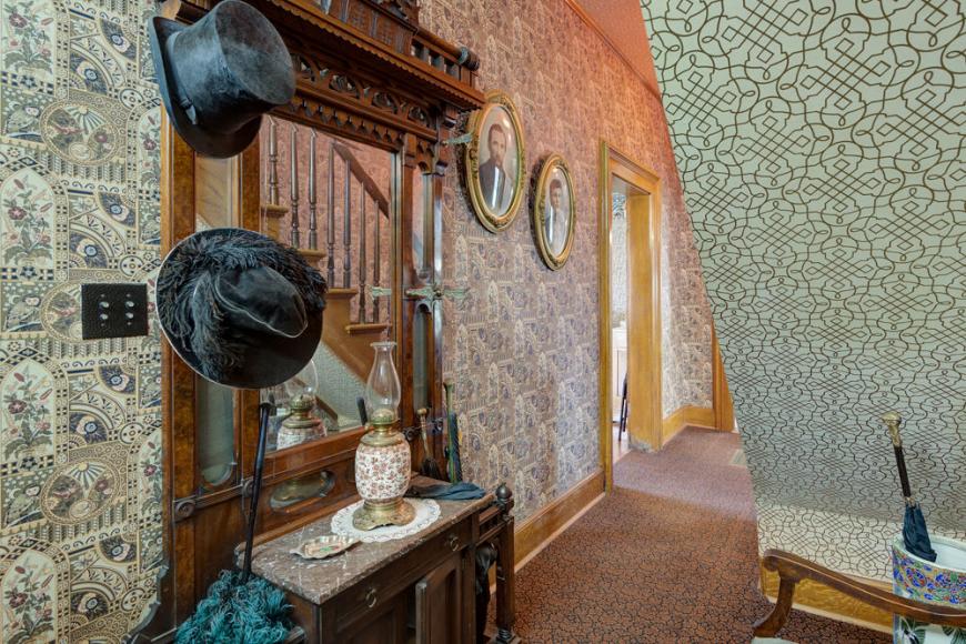 An entry way under a staircase reflected in a Victorian mirror and sideboard holding top hats.