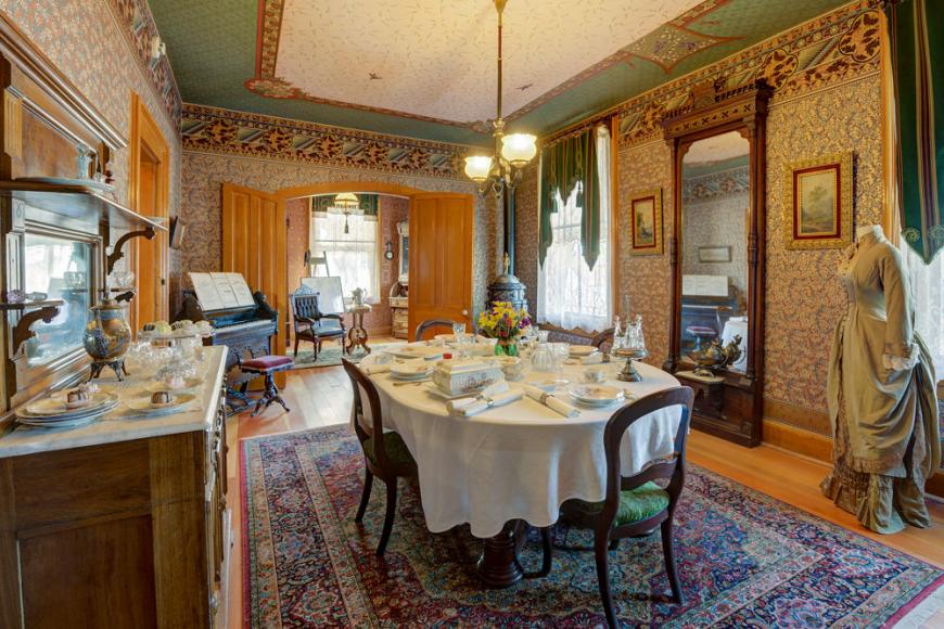 A heavily furnished Victorian dining room featuring a set table with a white tablecloth, sideboard and full length mirror.