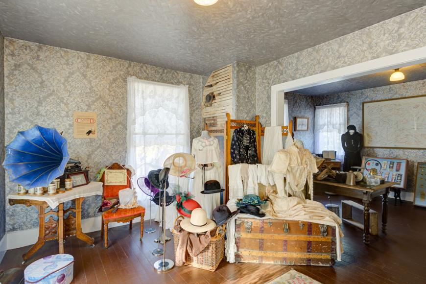 A grey wallpapered room featuring a display of various linens and textiles, a wooden chest and a bright blue gramophone.