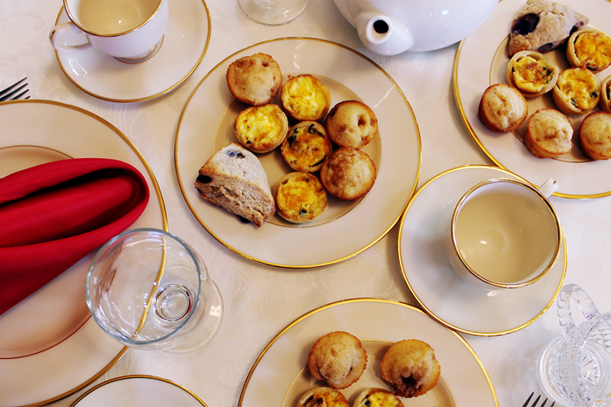 An overhead shot of multiple plates on a white tablecloth. They are filled with quiches, scones, and muffins and surrounded by tableware and teacups.
