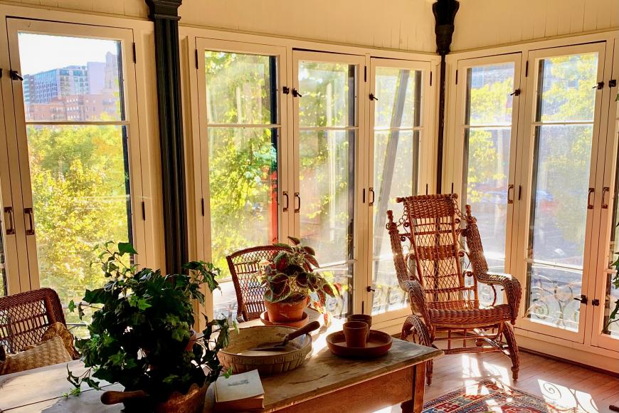 Interior of Center for Colorado Women's History sun room. Wooden rocking chairs sit in a room full of windows with lush greenery showing from outside.