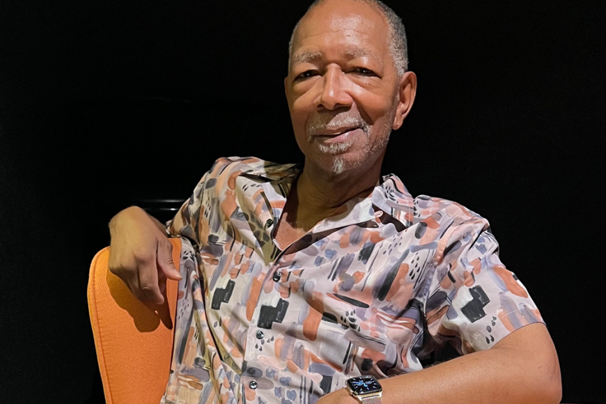 Ron Ivory in a patterned short sleeved shirt leaning against a chair and smiling. 