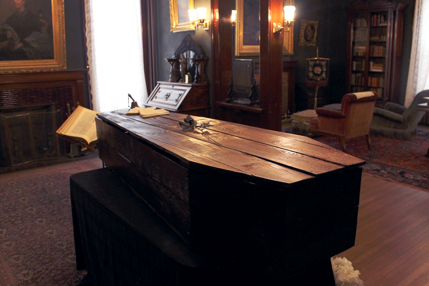 View of the Center for Colorado Women's History library. At the center of the room is a dark wooden coffin. On the lid is a black rose and a bell next to a small journal.