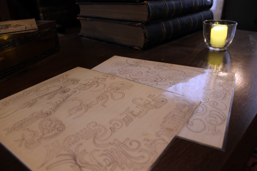 Dark wooden table lit with a small candle. On the table are sheets of automatic writing filled with thin, curving sloped shapes.