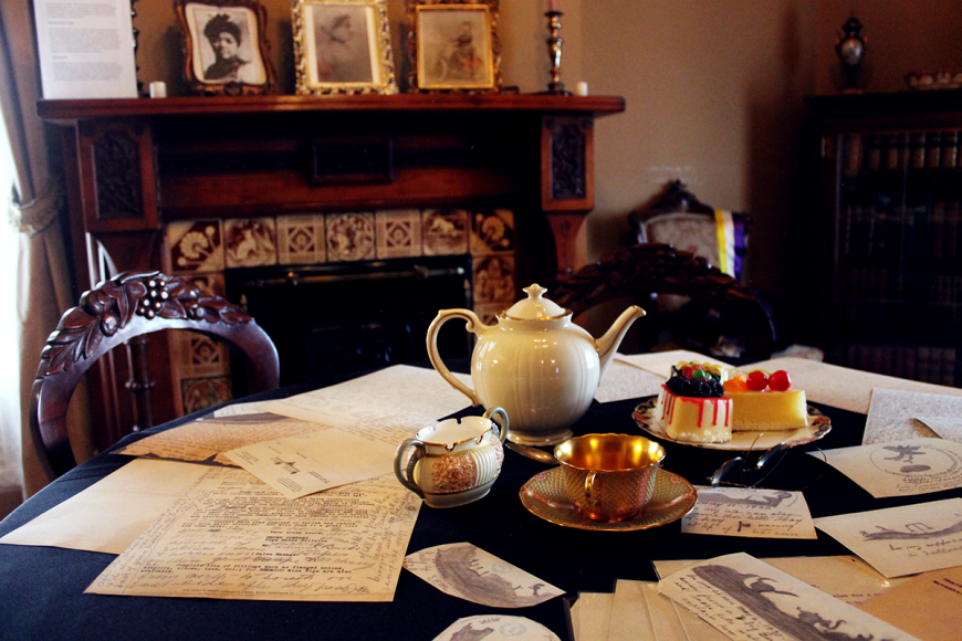 Ornate wooden table with a tea set and fruit tarts. Surrounding the tea setting are automatic drawings and tea leaf analysis drawings strewn across the table.