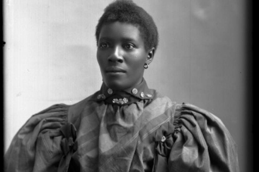 An African American woman in a victorian dress with puffy sleeves stares with a dignified expression at the camera.