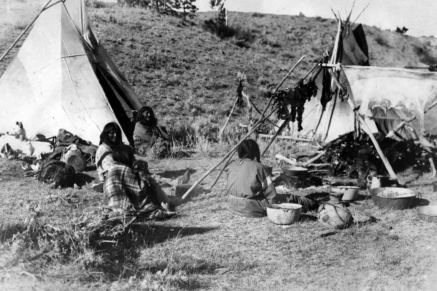 Ute women sitting outside 2 tepees and a hearth with hanging deer meet and hide hanging off of it.