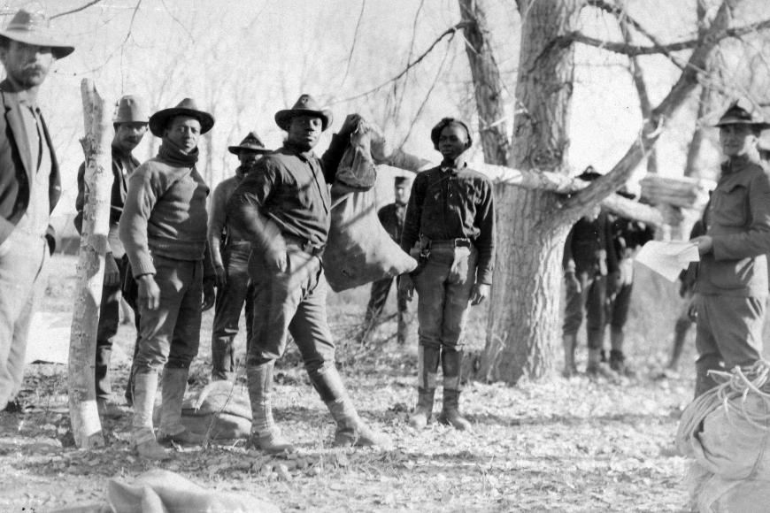 3 buffalo soldiers in military garb facing the camera. The man at the center holds a large burlap sack. Around them are white men in military and formal dress looking at the camera.