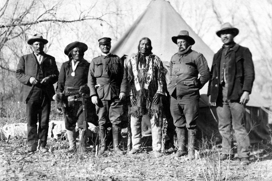 Officers, Interpreters, and Sioux Diplomats standing in a line facing the camera. They wear military garb before a tepee.