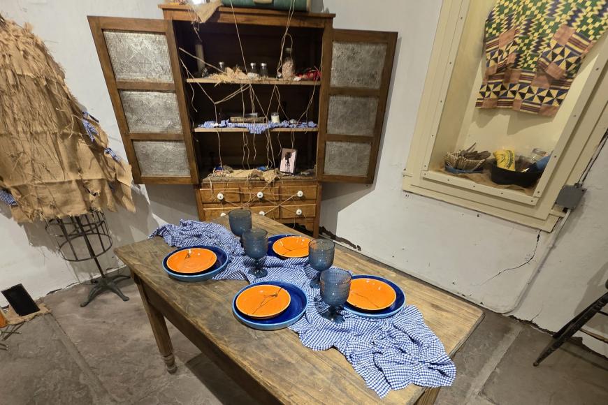 Historic table setting and cabinet covered in colorful, artistic tableware and artifacts, part of the 12 tablecloths exhibition. Behind the table is a dress made of burlap.