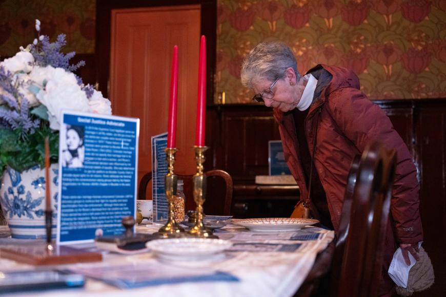 A female guest leans over the antique dining table covered in artifacts and an informational panel.