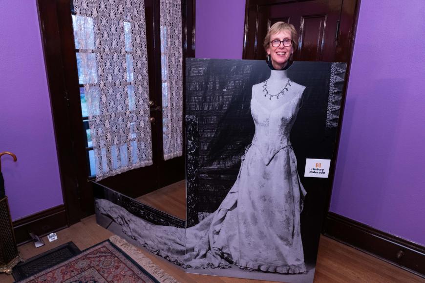 A museum guest in front of a purple background posing with a cardboard cutout of Baby Doe Tabor's wedding dress below her neck.