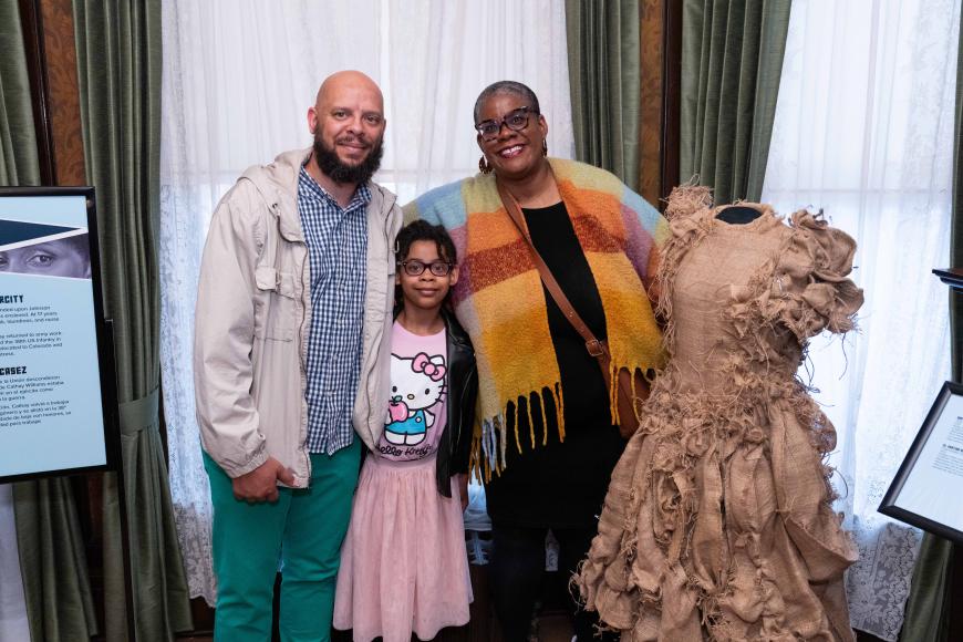 Artist Chloe Duplessis stands with her partner and daughter next to her art piece, Sista Soldier, an ornate burlap dress with bows and braids embedded through it.