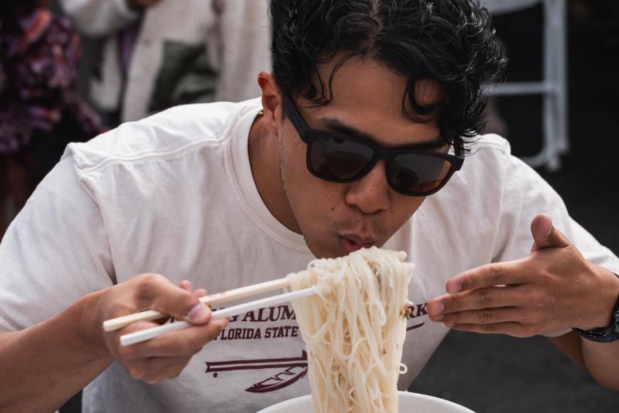 A man in sunglasses eats a large bowl of noodles with a pair of chopsticks.