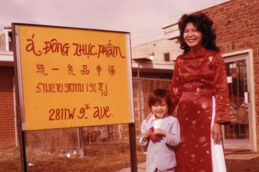 A mother and child in Vietnamese traditional clothing pose near a sign reading: á đơng Thực Phẩm