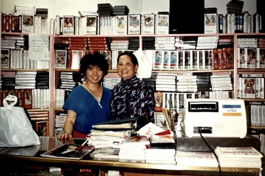 2 women smile behind the counter which is fully stocked with VHS rentals.