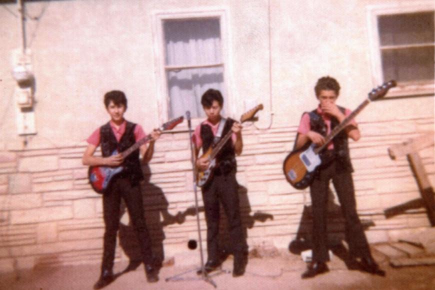 3 young men post in matching pink shirts and black vests. They each hold an electric guitar and pose for their performance.