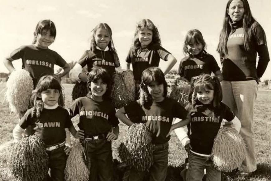 8 cheerleaders and 1 adult trainer post with their hands on their hips holding pom poms. They each wear shirts with their first names on it.