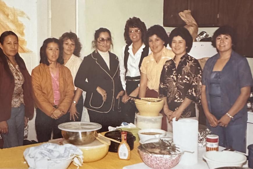 A group of mothers stand before a table of food they have prepared for a community event.