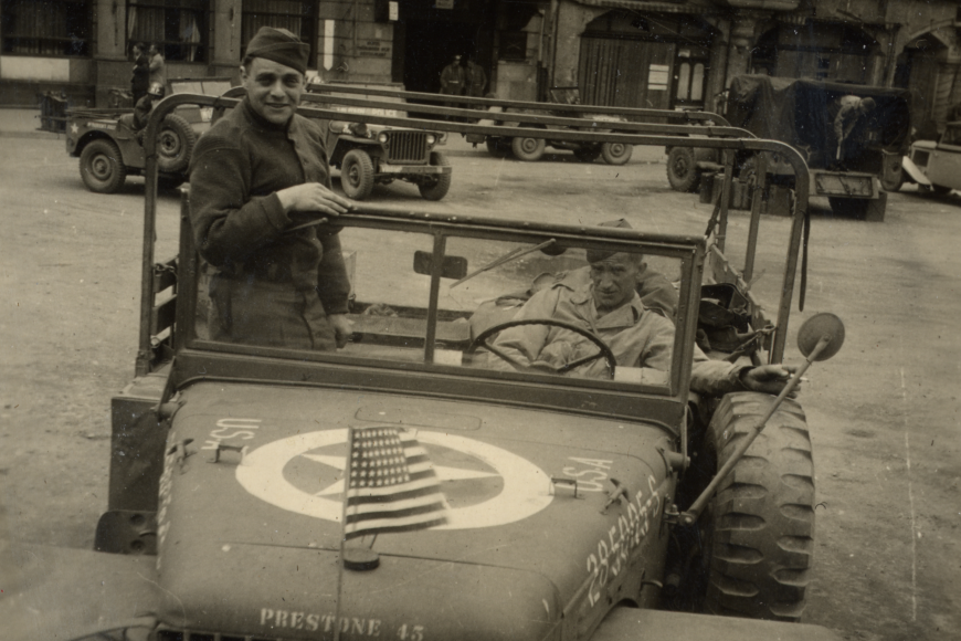 Joseph Eaton in uniform, stand in his open top military jeep and smiles at the camera.