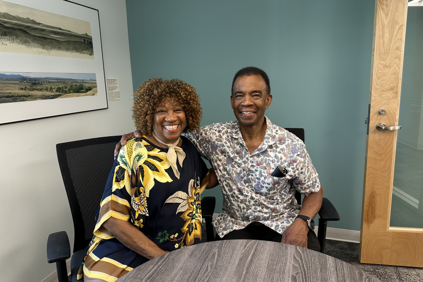 2 adults sit side by side at a round table and look at the camera smiling.