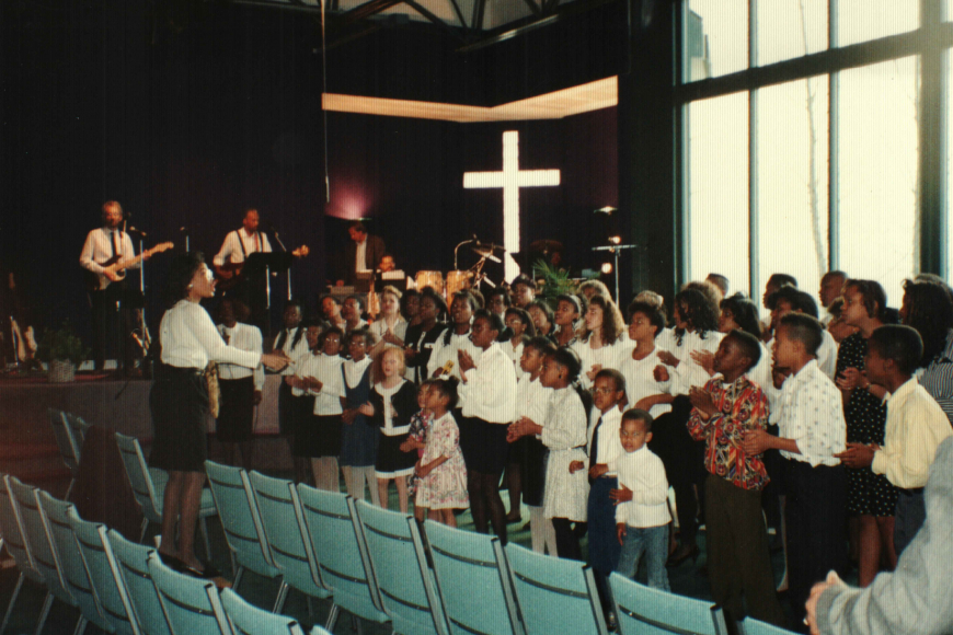 3 rows of children of all ages sing in a church choir with a band to the right of them.