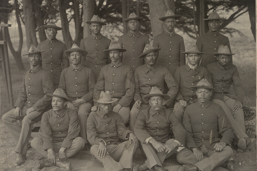 A group of Black Buffalo Soldiers stand in rows and face the camera in uniform.