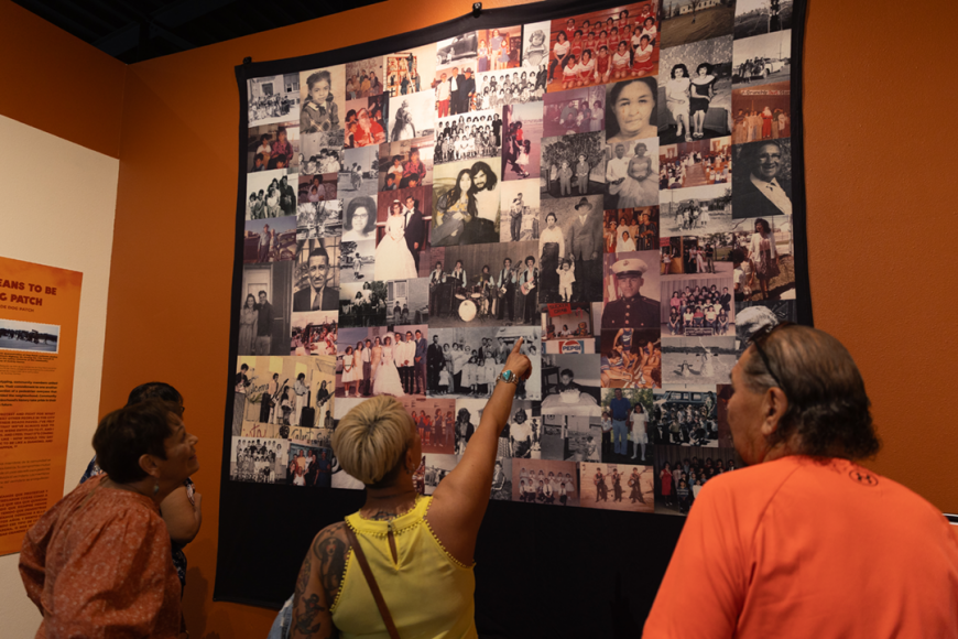 A group of 3 visitors point at the Dog Patch Quilt of community photos hanging on a wall.