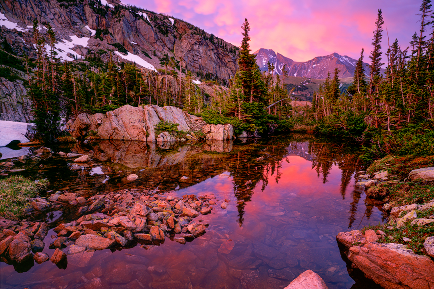 Long’s Peak at Sunrise with a reflective lake casting a pink and purple glow across the image.