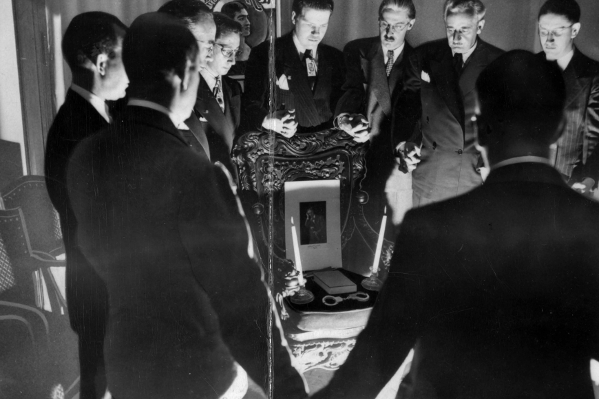 A group of men stand in a circle holding hands. There's a ghostly light shining on their faces from below.