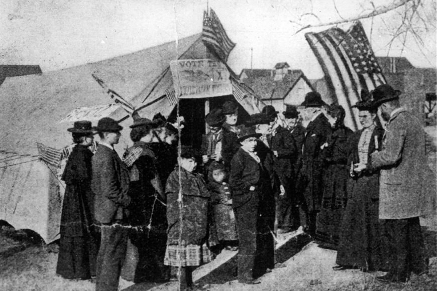 A group of men and women stand outside a precinct headquarters tent with banners and American flags. 